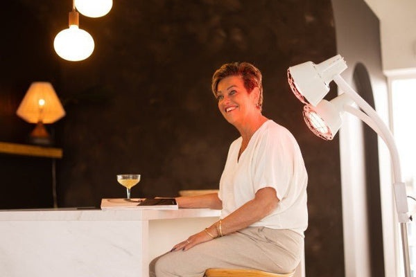 Lachende vrouw aan tafel met naast zich een staande infraroodlamp met twee warmtelampen