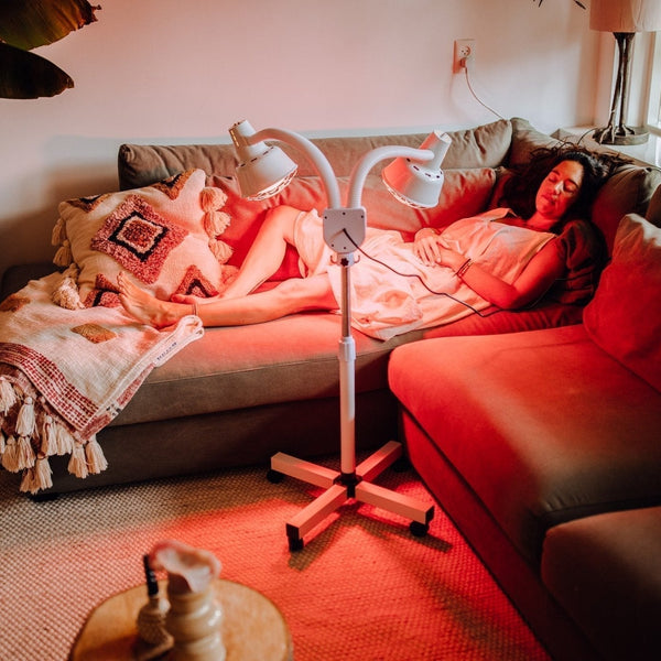 Vrouw ligt ontspannen op de bank onder een infraroodlamp met twee koppen op een rolstatief
