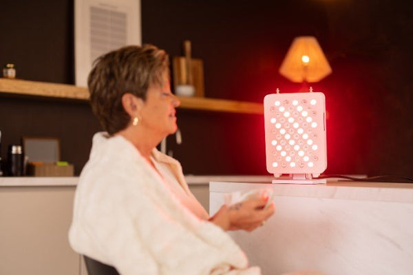 Vrouw in badjas met een mok zit voor een klein rood licht therapie paneel dat haar gezicht belicht